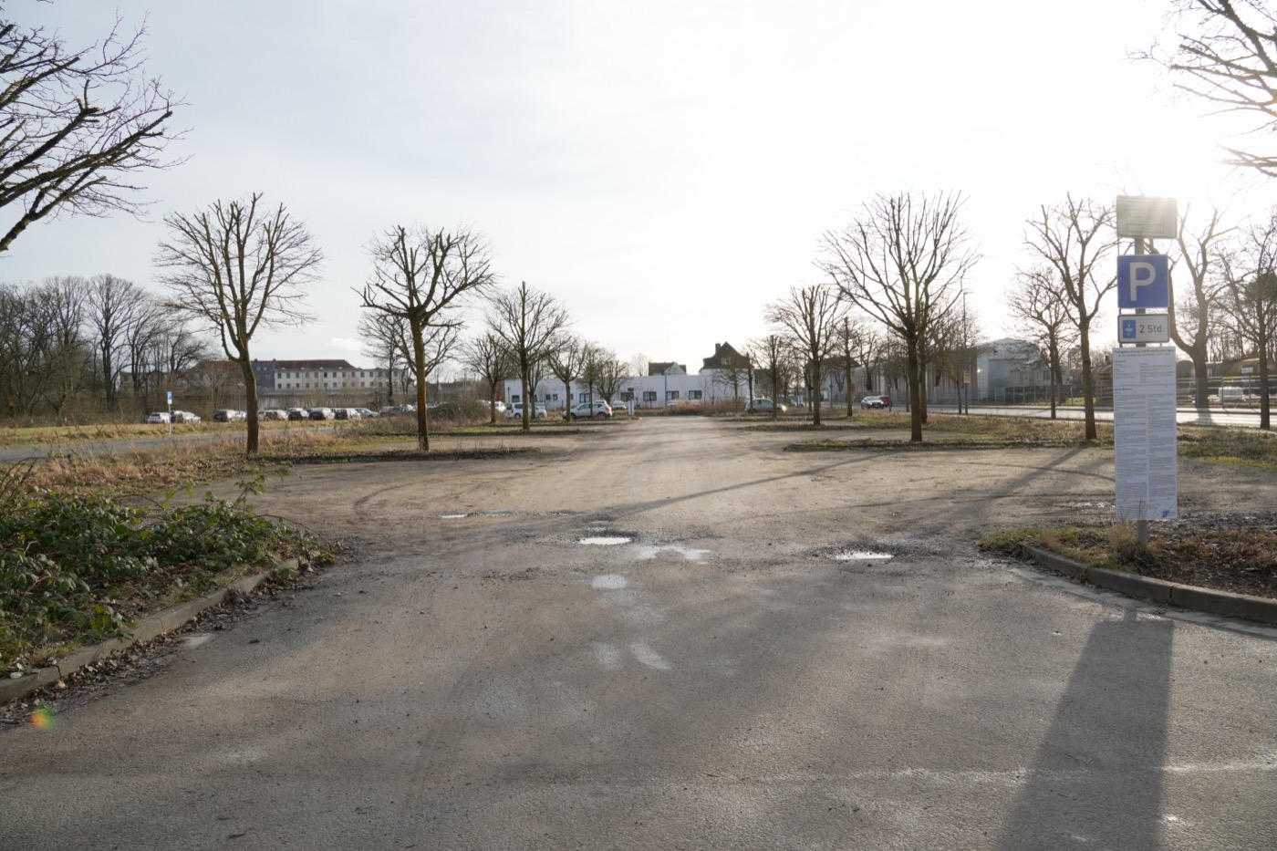 Großer Parkplatz mit leeren Parkbuchten, Bäumen und einem Schild an der EInfahrt.
